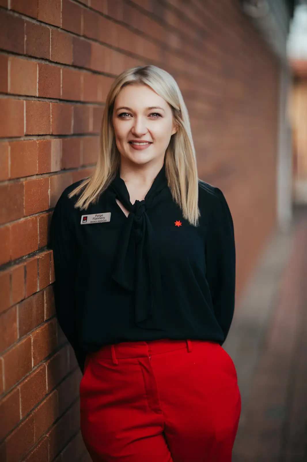 Branch manager leaning on a brick wall, black top and red trousers