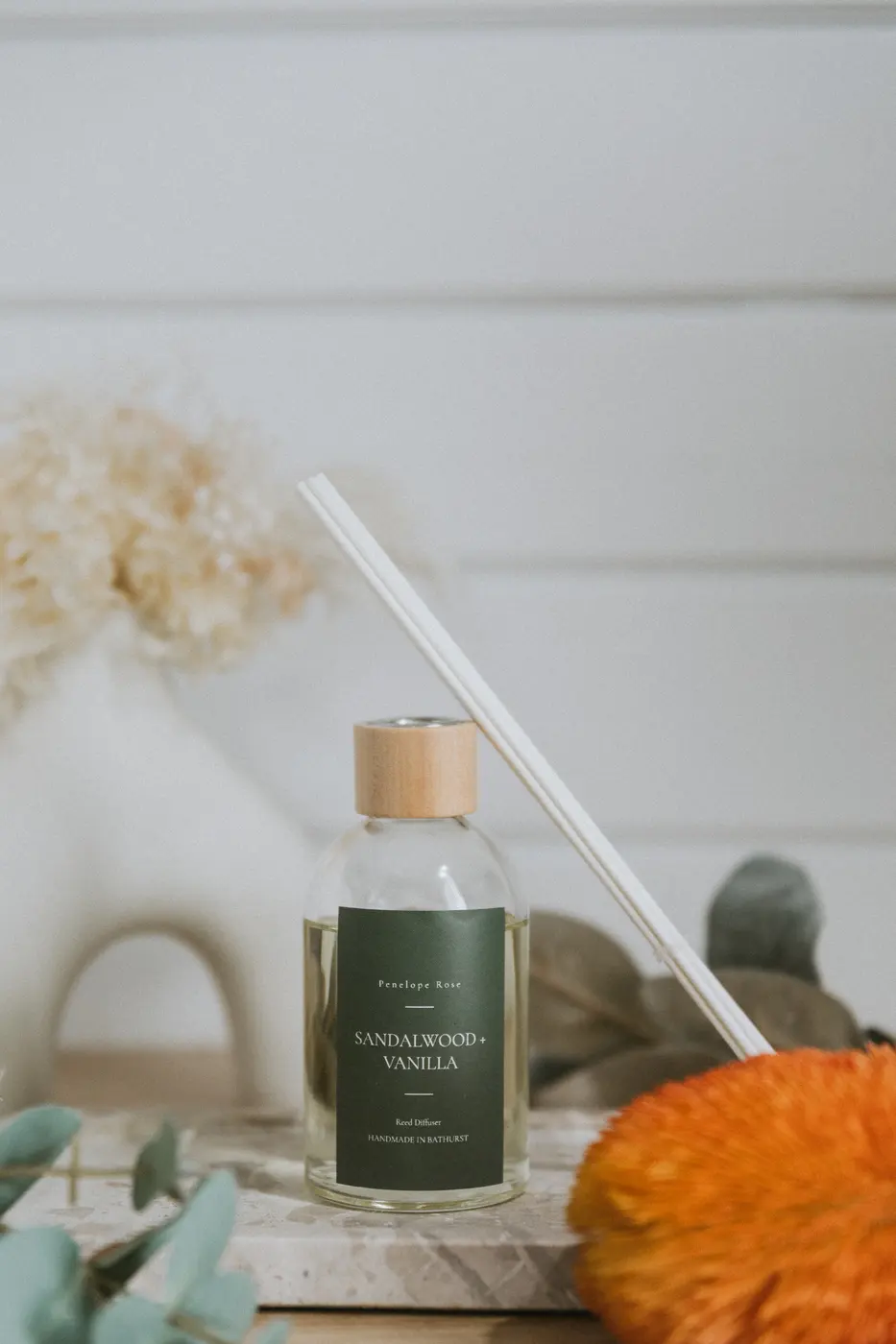 Glass reed diffuser with an upright reed and green label, soft-focus florals behind
